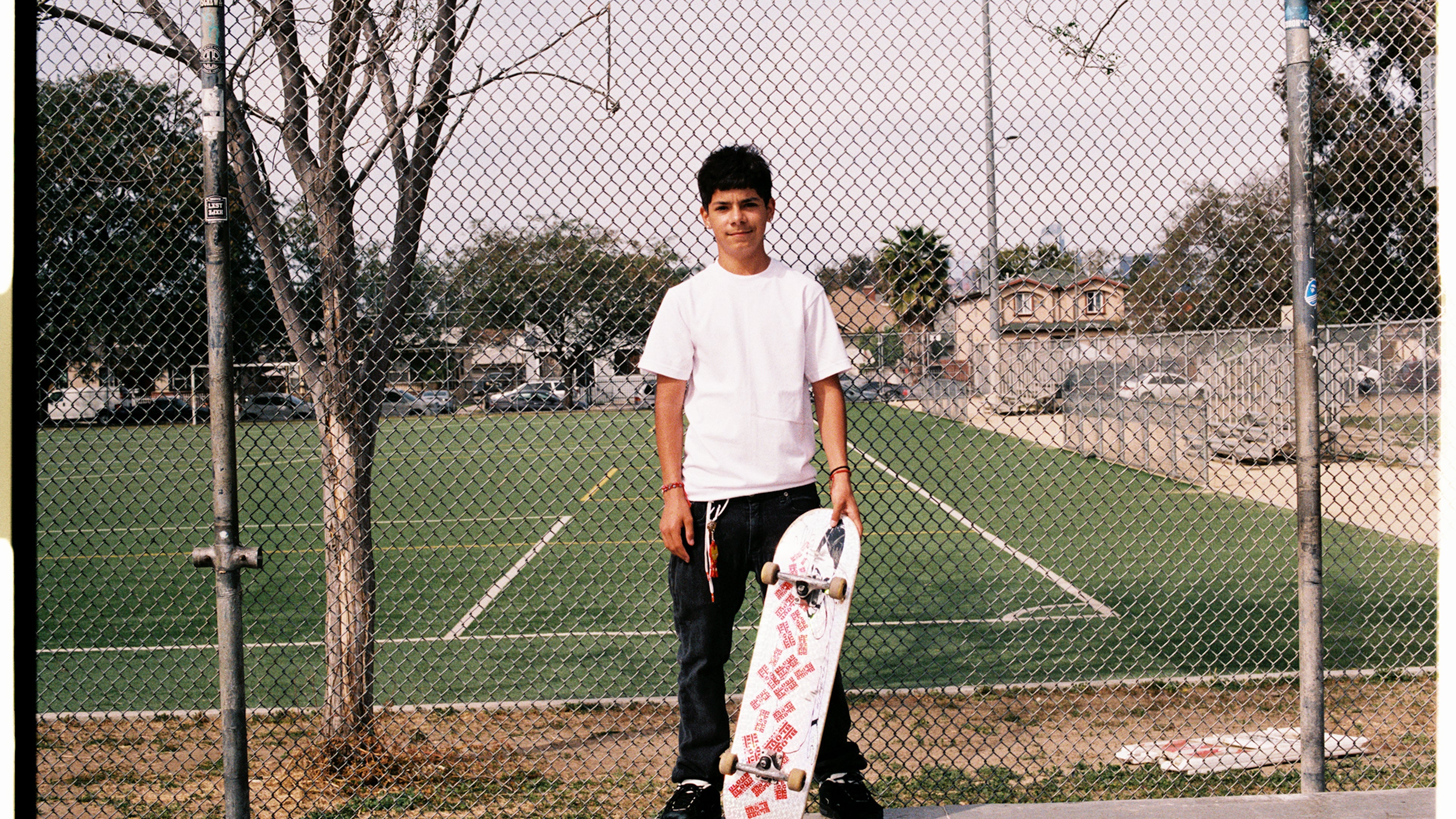 Young Hispanic male is holding a skateboard wearing a Shaka Wear T shirt in front of a field.