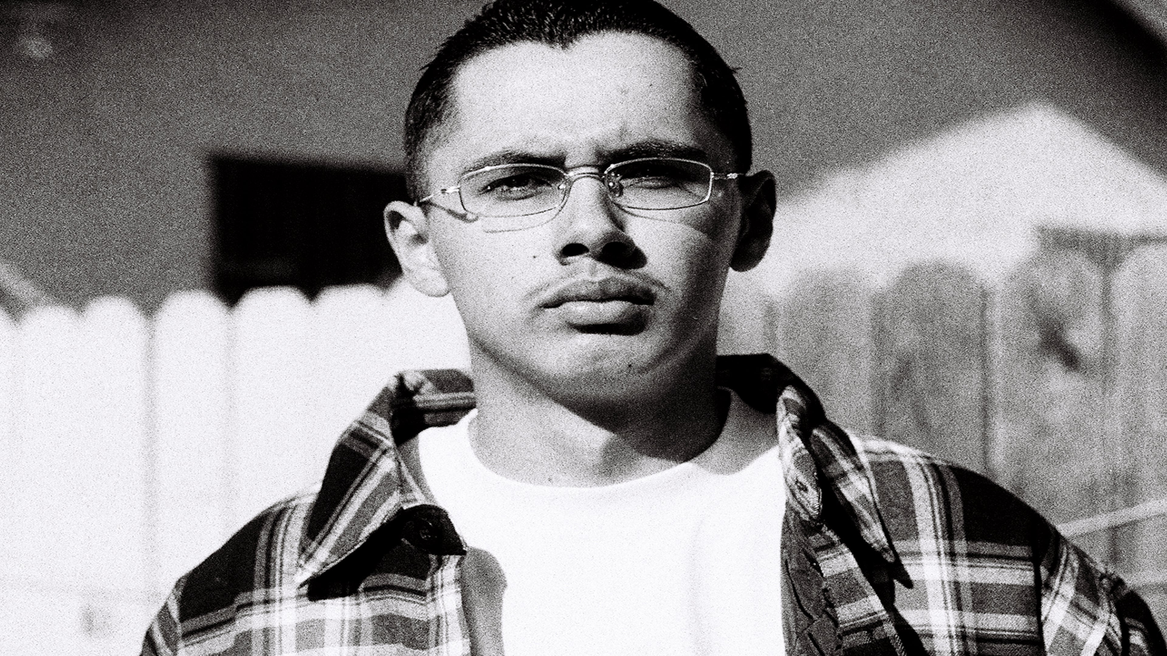 A hispanic man stands in front of a house wearing a Shaka Wear Flannel over his White T shirt.