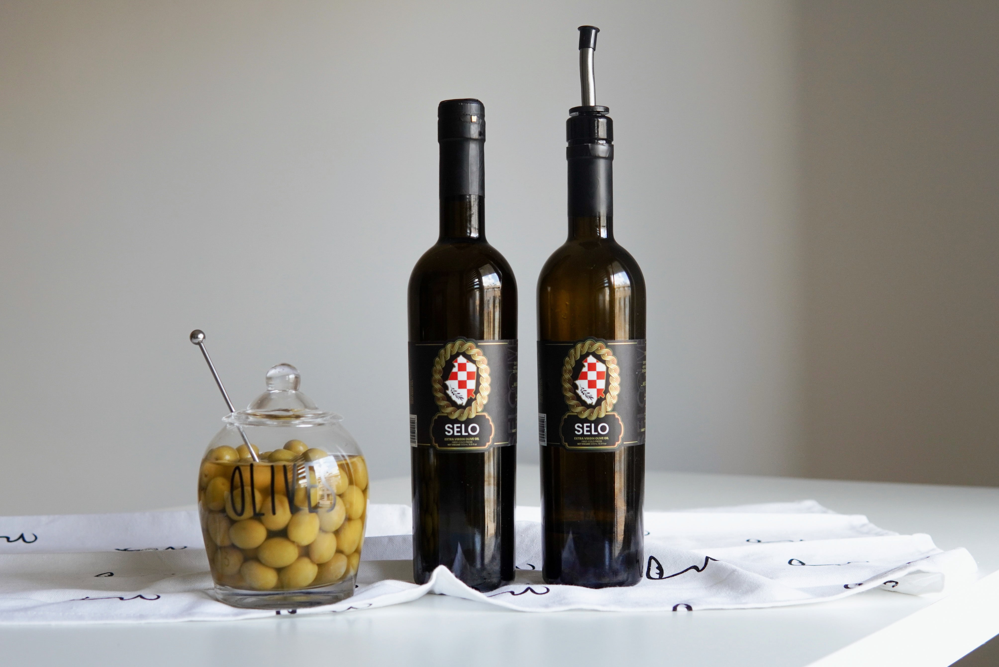 Elegant Selo olive oil bottles and a jar of olives on a white tablecloth, creating a sophisticated and minimalist presentation.