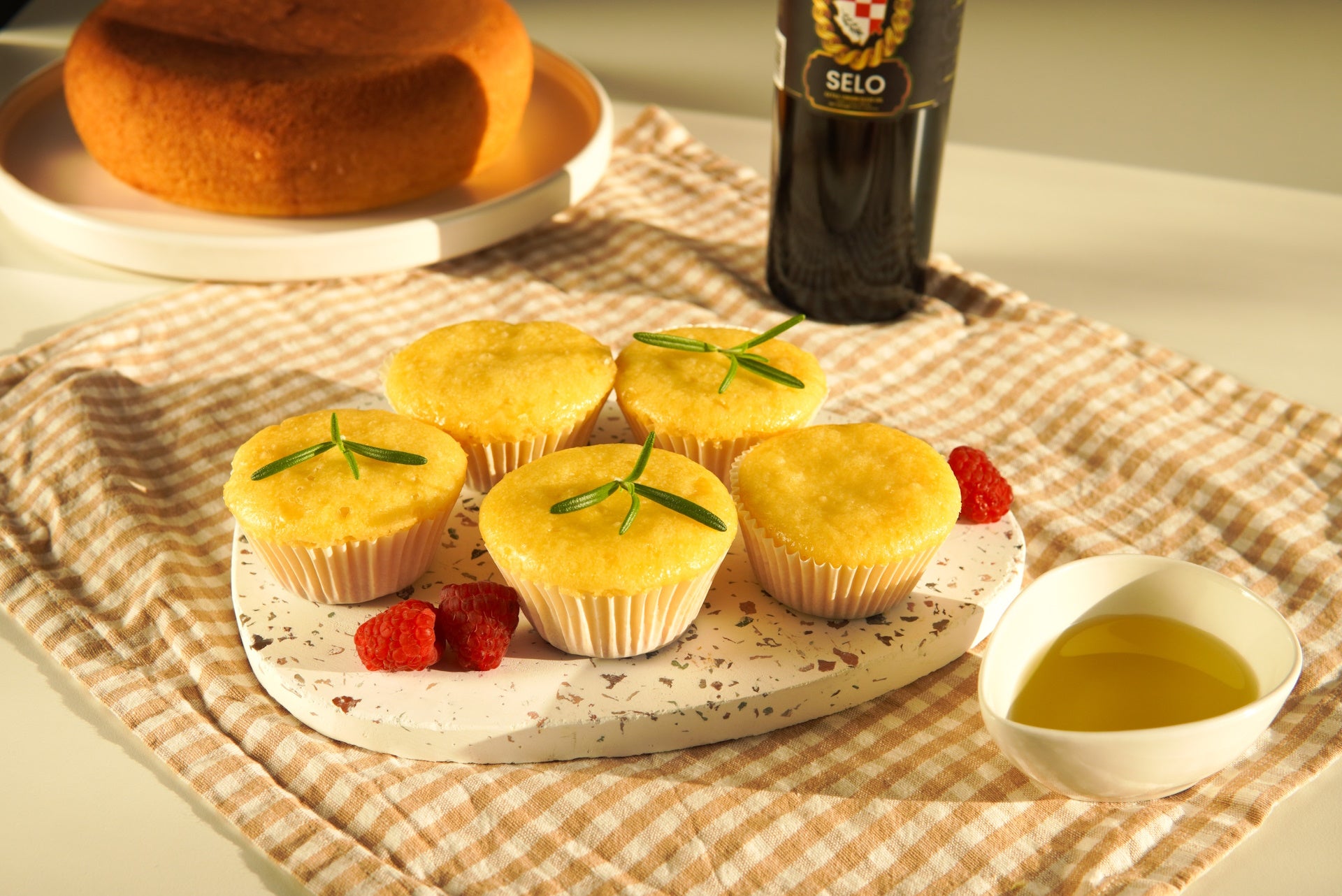 Image of a batch of fluffy cupcakes baked using Selo Croatian extra virgin olive oil, beautifully arranged on a pastel cupcake stand, with a clear glass bottle of Selo olive oil tastefully positioned in the background.