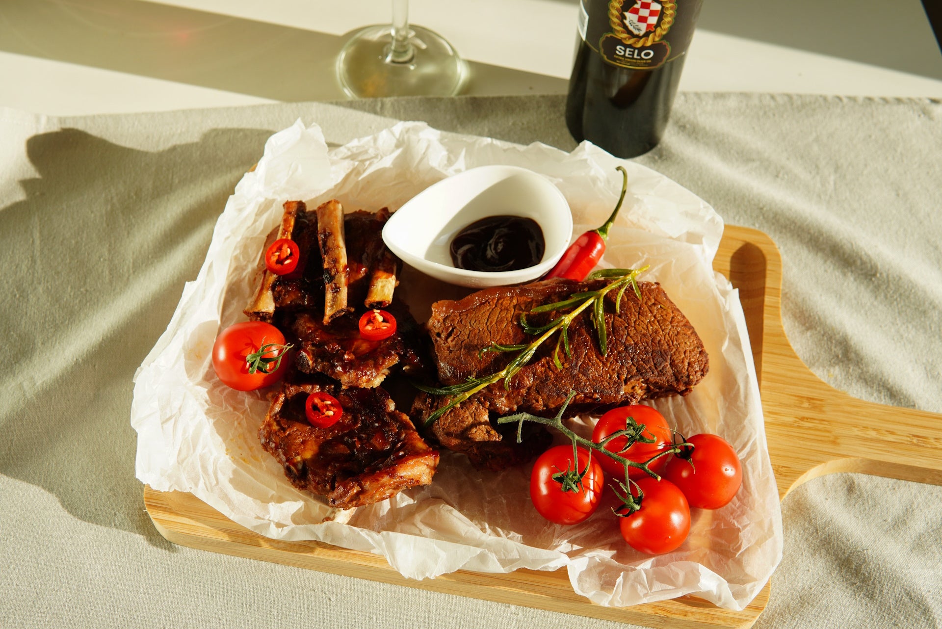 A plate of grilled meats, lamb ribs, tomatoes, and peppers, accompanied by a bottle of Selo olive oil and a glass of red wine.