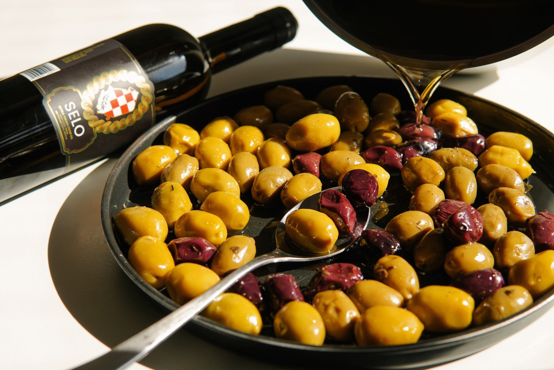 Close-up of a bottle of Selo Croatian extra virgin olive oil next to a plate of green and black olives, with olive oil being poured over them.