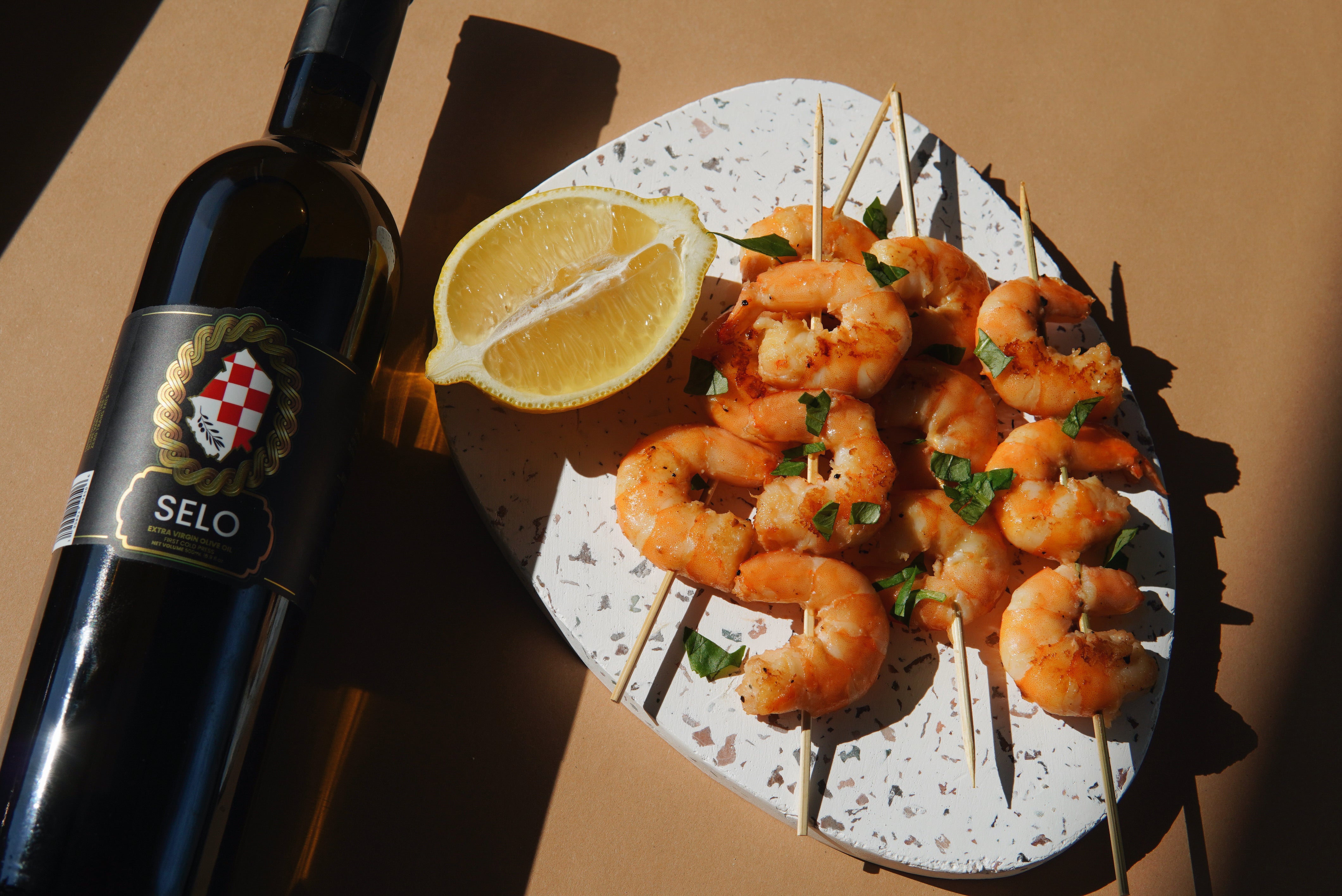 A bottle of Selo Croatian Extra Virgin Olive Oil stands elegantly next to succulent grilled shrimp, their pink hues beautifully complemented by the rich golden color of the olive oil.