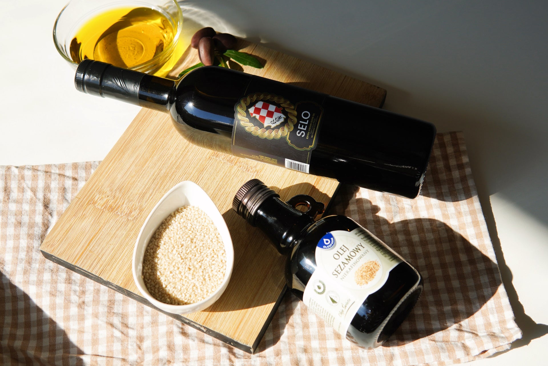 On a beautifully spread tablecloth over a marble surface, two sleek black bottles make their presence — sesame oil and olive oil. Adjacently, a bowl filled with fresh sesame seeds paints a picture of nature's simplicity and elegance.