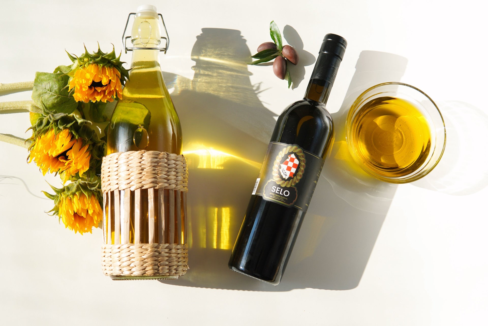 On a flawless marble countertop, a bottle of sunflower seed oil stands next to an olive oil bottle; their contrasting golden and green tints, paired with elegant designs, represent the diverse richness of nature and artful craftsmanship.