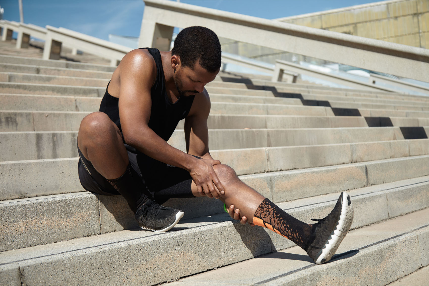 Handsome black runner with muscular athletic body holding his leg with both hands, feeling pain in knee or calf, massaging it, suffering from strain or spasm while sitting on steps of concrete stair