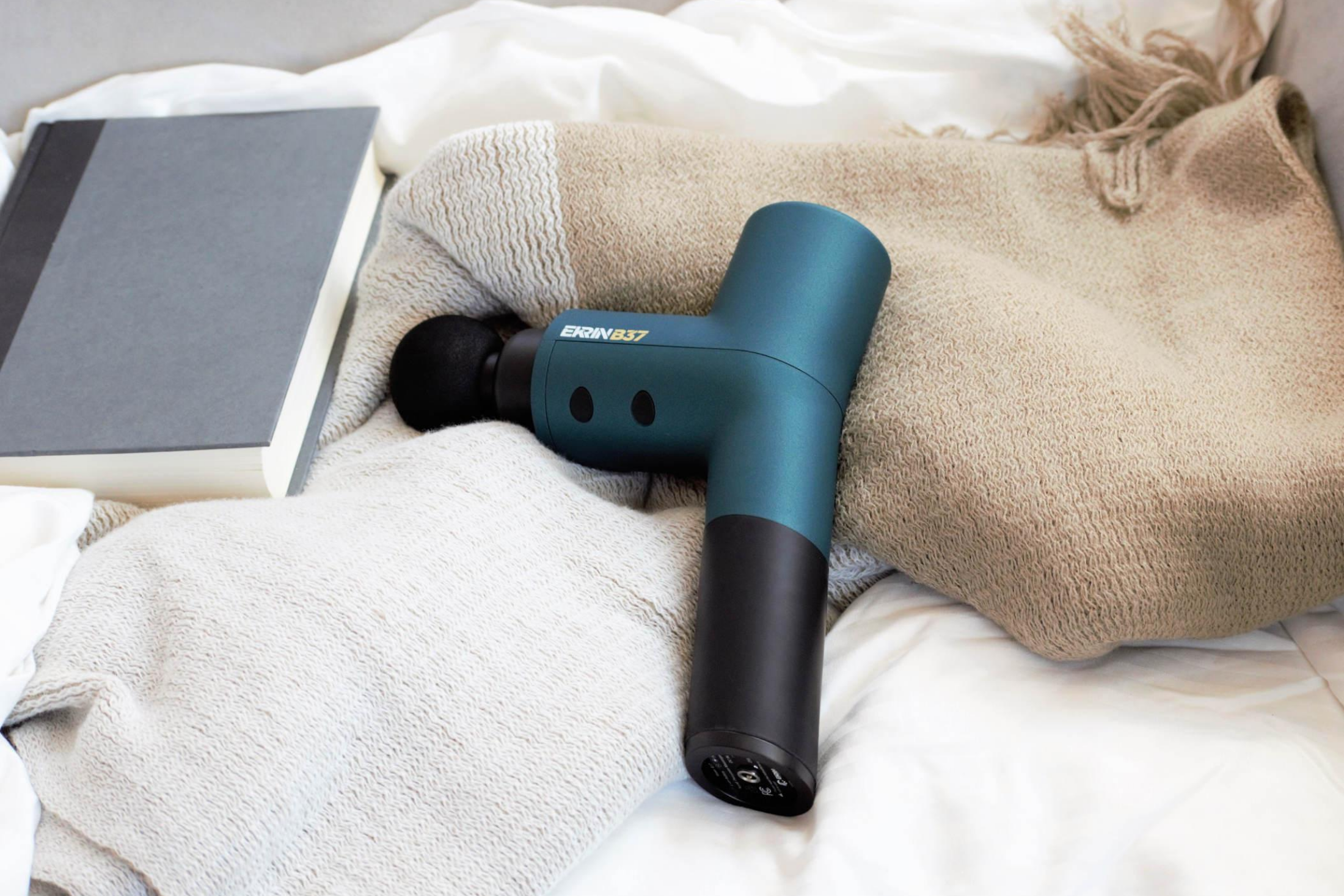 Ekrin B37 massage gun on a bed next to a book and blanket