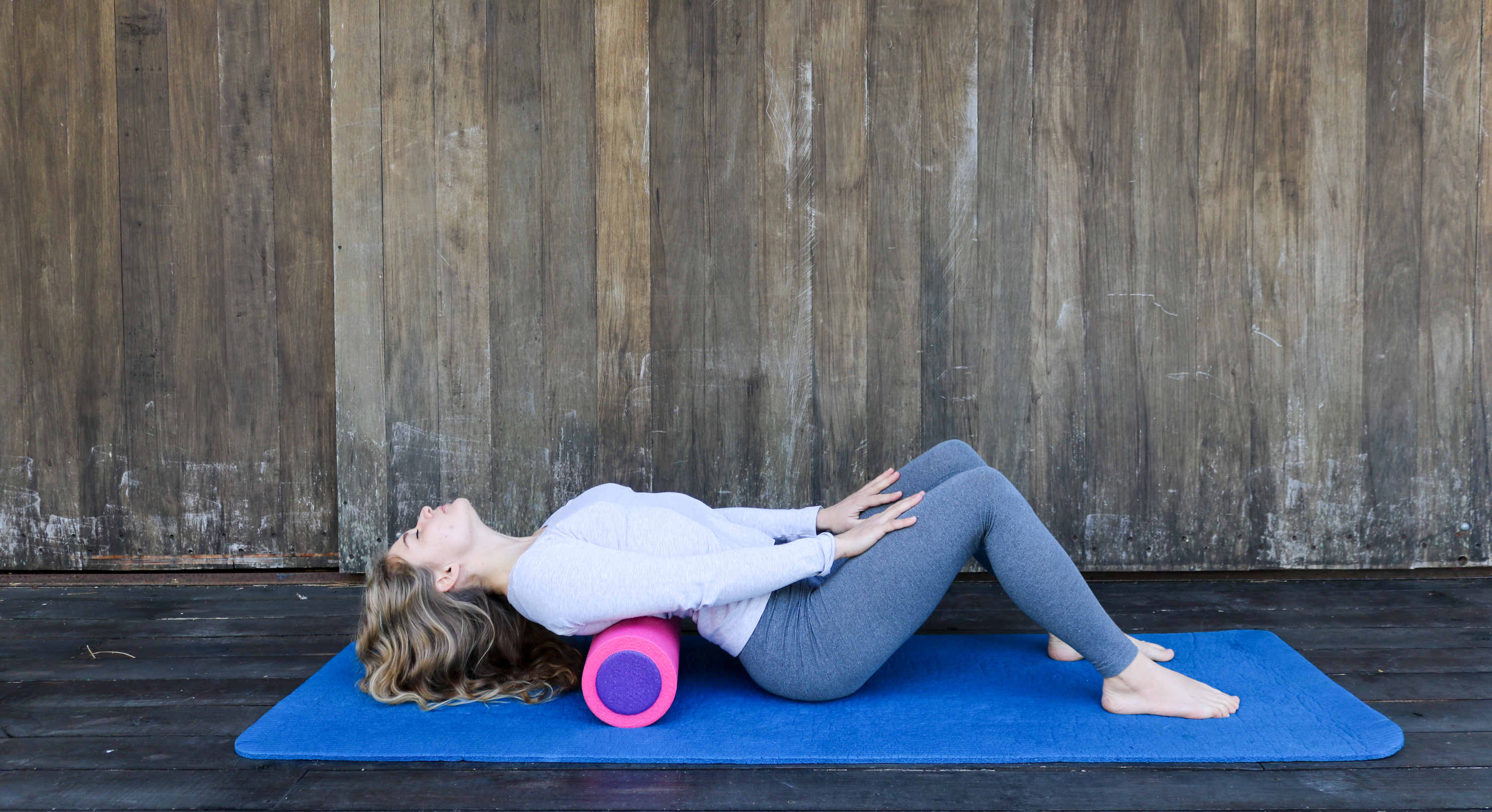 A young blond caucasian woman rolling her back on a foam roller on a blue yoga mat, wooden floor and wooden background.