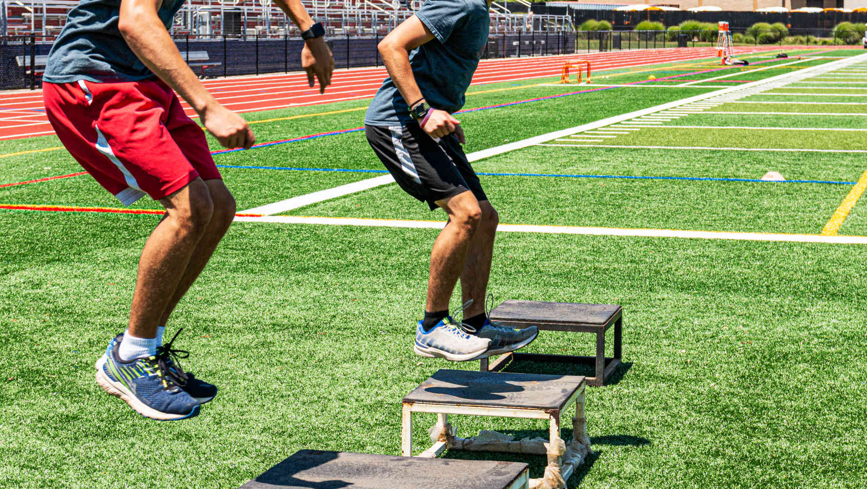 Two male high school track and field athletes doing box jumps on a green turf field during strength and agility practice.