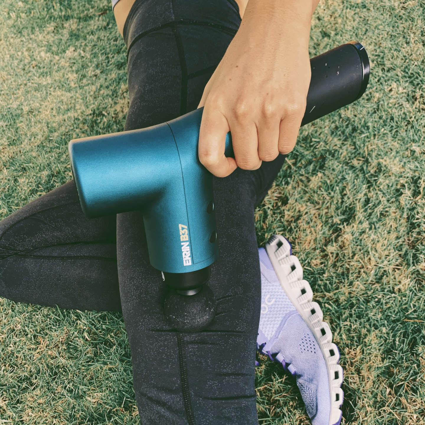 Athletic woman massaging her muscles with a massage gun outside on grass