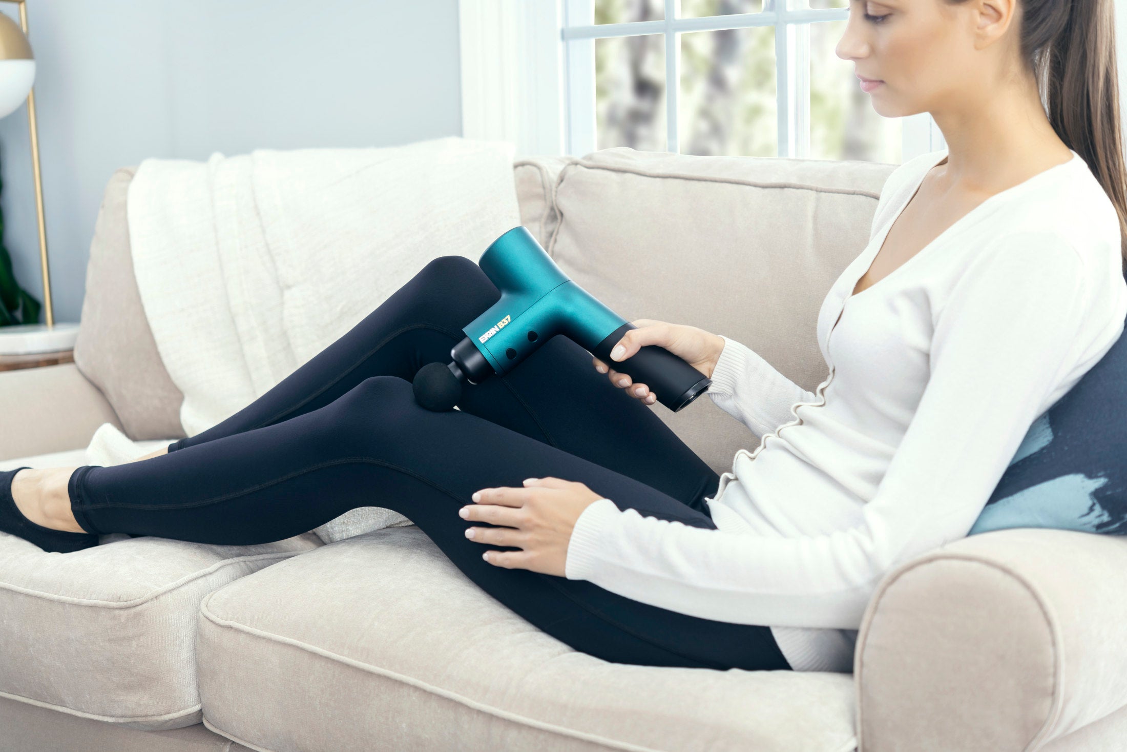 Athletic woman using Ekrin B37 massage gun on a white couch