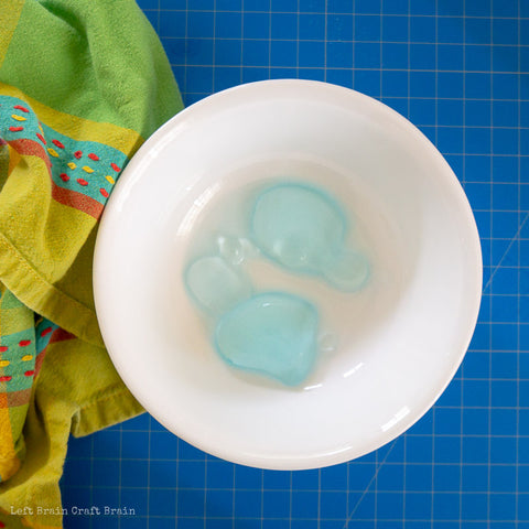 A white bowl holds water and several edible water bubbles. There is a kitchen towel next to the bowl on a blue counter. Credit to Left Brain Craft Brain