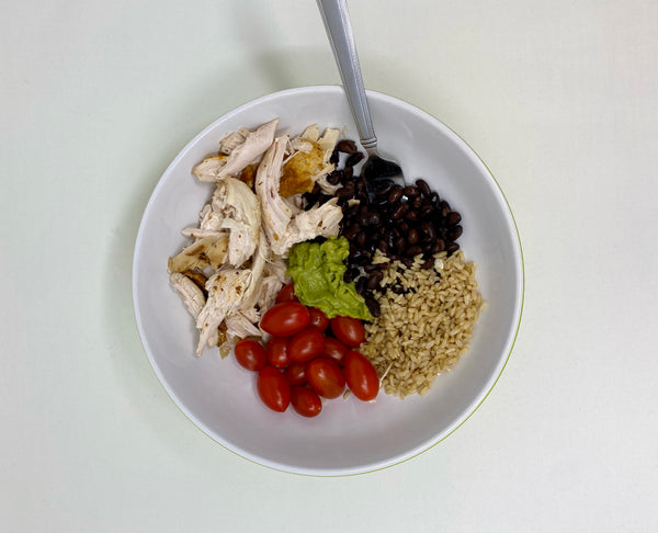 : on a plate is sliced rotisserie chicken, tomatoes, black beans, brown rice and a spoonful of guacamole in the middle.