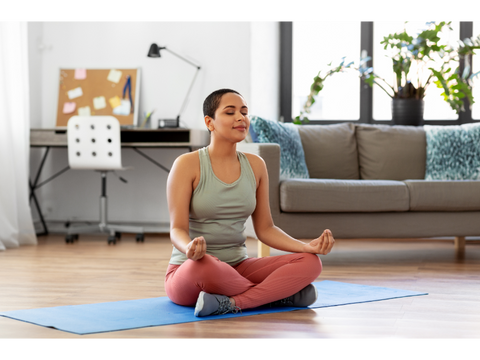 Woman sitting cross-legged on a yoga mat in her living room while meditating.