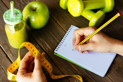 A person writing down a list of to dos on a pad of paper. The pad is on a table with a healthy smoothy, an apple, and weights.