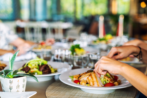 Hands cutting into chicken on top of a salad in a white bowl. There are other bowls of healthy food on the table in a restaurant.