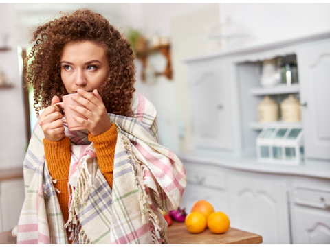 woman in her kitchen with a blanket over her shoulders clutching a warm cup of tea