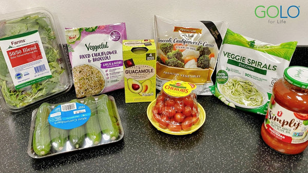 plastic container of 50/50 blend salad mix, package of five mini cucumbers, bag of rice cauliflower and broccoli, box of pre-packaged guacamole, package of cherry tomatoes, package of frozen broccoli and cauliflower, package of zucchini veggie spirals, and a jar of Simply brand marinera sauce on a counter top.