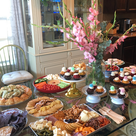 a glass dining room table set up buffet style with crudite, cupcakes, and other party snacks with a looming bouquet of pink gladiolas in the center