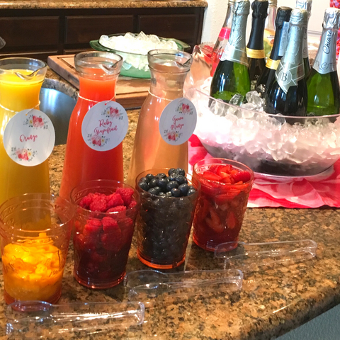 a diy mimosa bar with three carafes of different colored juice and cups of sliced fruit on the left and a large bowl full of ice and wine bottles on the right