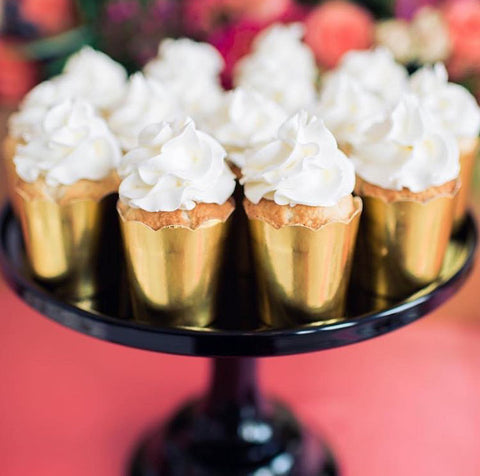 a red table top with a glossy black cake stand holding a dozen individual mini cakes in gold foil wrappers topped with whipped cream