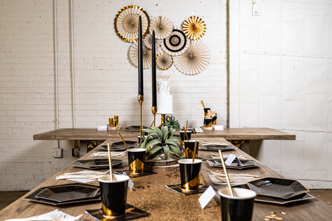 a long head table dressed with black and gold plates and cups for a party, with a set of black, white, and gold party fans on the wall behind them