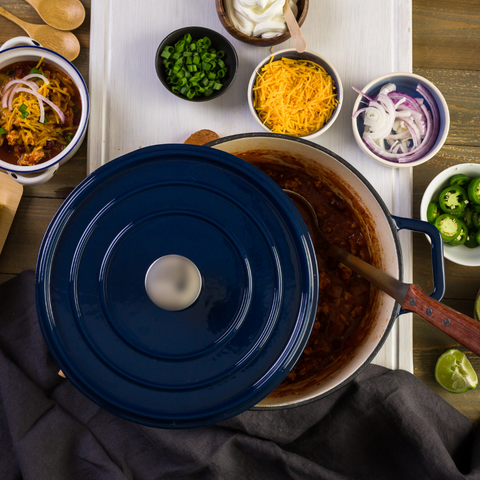 A large blue cast iron pot view from above with the lid half off to show chili inside. Around the pot in a semi circle is an assortment of toppings in small bowls like cheese and onions