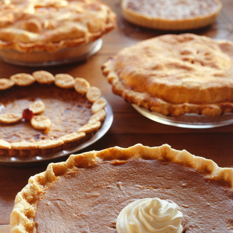A whip cream topped sweet potato pie sits in the foreground with several other topped and decorative crust pies behind it on a wooden table