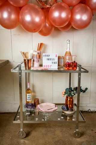 a metal bar cart styled with copper cups, Eucalyptus, and gem stones- with rose' bottles and a letter board that reads " stop and smell the rose' "