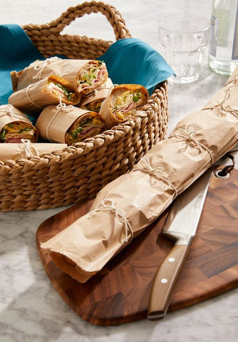 a basket on a grey countertop full of sub sandwiches wrapped in brown paper, sitting behind a cutting board  with one long, uncut wrapped sandwich and knife on it