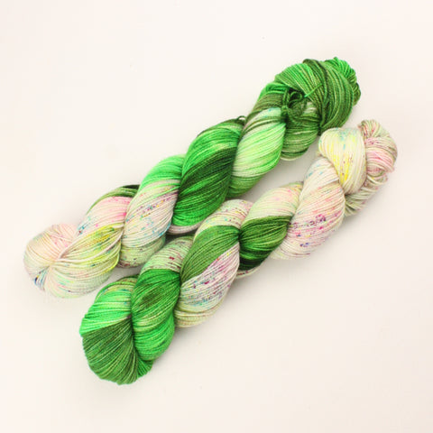 two skeins of yarn laying on a white background. their colours start at all colours of leafy green at one end. melting into neon cake like speckles at the other end 