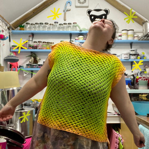 a photo of me standing in my dye shed wearing a giant crochet square top which starts with a lime green in the middle and transitions to light pink via yellow. i am standing with my hands out to my side looking up to the roof with a ridiclous look on my face