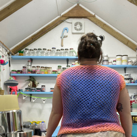 a photo of me standing in my dye shed but with my back to the camera to show off the back of my giant granny square top. the back of the top is exactly the same as the front but its fades from an indigo purple through to pink. 