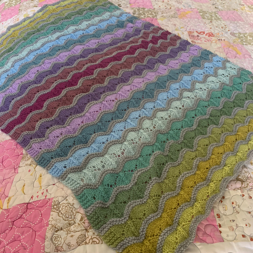 bounce baby blanket laying on top of a pink and white quilted blanket. the blanket has a wave pattern to it with each pattern repeat being a different colour seperated by a bump of light grey. the colours go through lime green to dark greens, light blues to dark blues light lilacs to dark purple and then back again to the lime greens. 