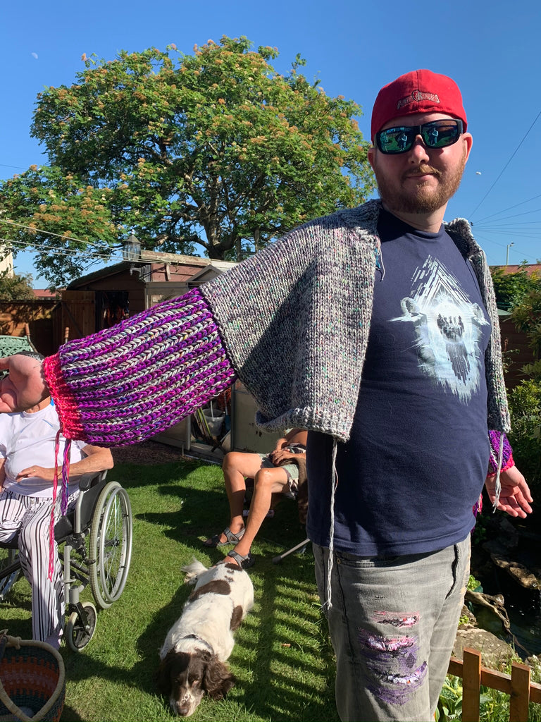 a man (mark) standing in a garden with a bright blue sky, a tree, a spaniel (oskar) and his grandparents in the background. he is wearing sunglasses a red powerrangers hat, a destiny 2 tshirt thats dark blue and the graffic looks like an iceberg with light denim jeans. the knit wear is the cardi bri that has a light grey main body and the sleeves are giant with purple and light grey brioche.