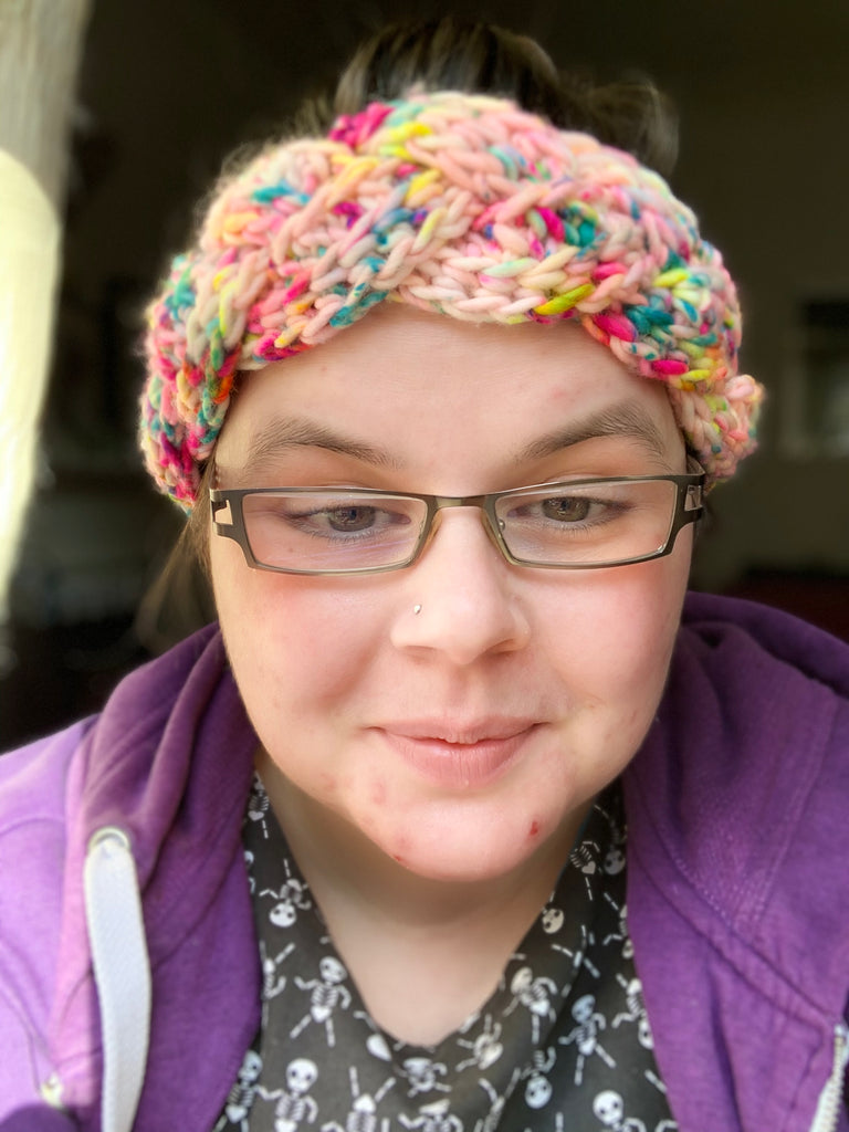 a really close up photo of my face. i have a black dress with skelletons on and a purple hoodie. im wearing grey rectangle glasses. i have on a crochet braided headband that is made from our atypical colourway, that colourway is a natural base with neon speckles of pink, yellow and blue
