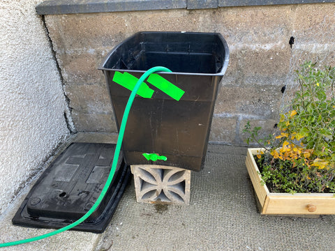 the main focus is a big black tub with neon green duct tape on the bottom and top covering some holes. a green hose is going into it. its being held up on some square decorative bricks and my tomato plant on the left