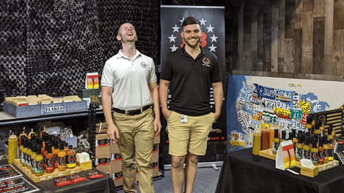 Sean and Eli smiling and laughing in front of their 13 Stars Hot Sauce booth