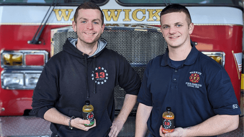 Sean from 13 Stars Hot Sauce holding a bottle of mean Green while standing in front of a firetruck and a firefighter