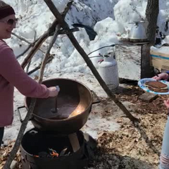 Fresh maple syrup being ladled from a copper kettle onto a plate of pancakes in a wintry woods