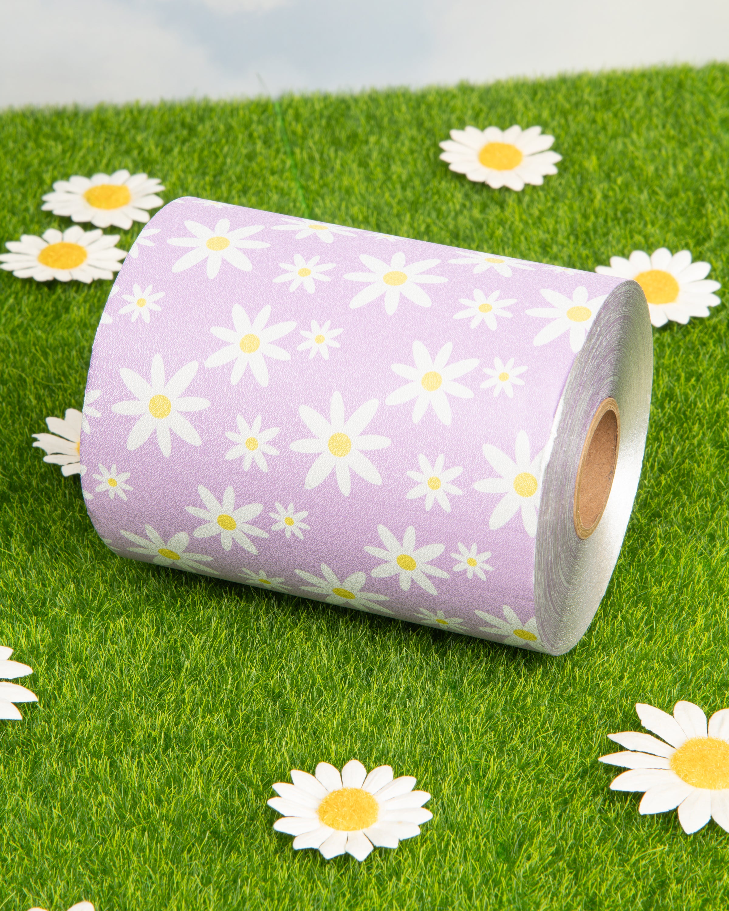 Roll of purple daisy-print toilet paper on artificial grass surrounded by white daisies.