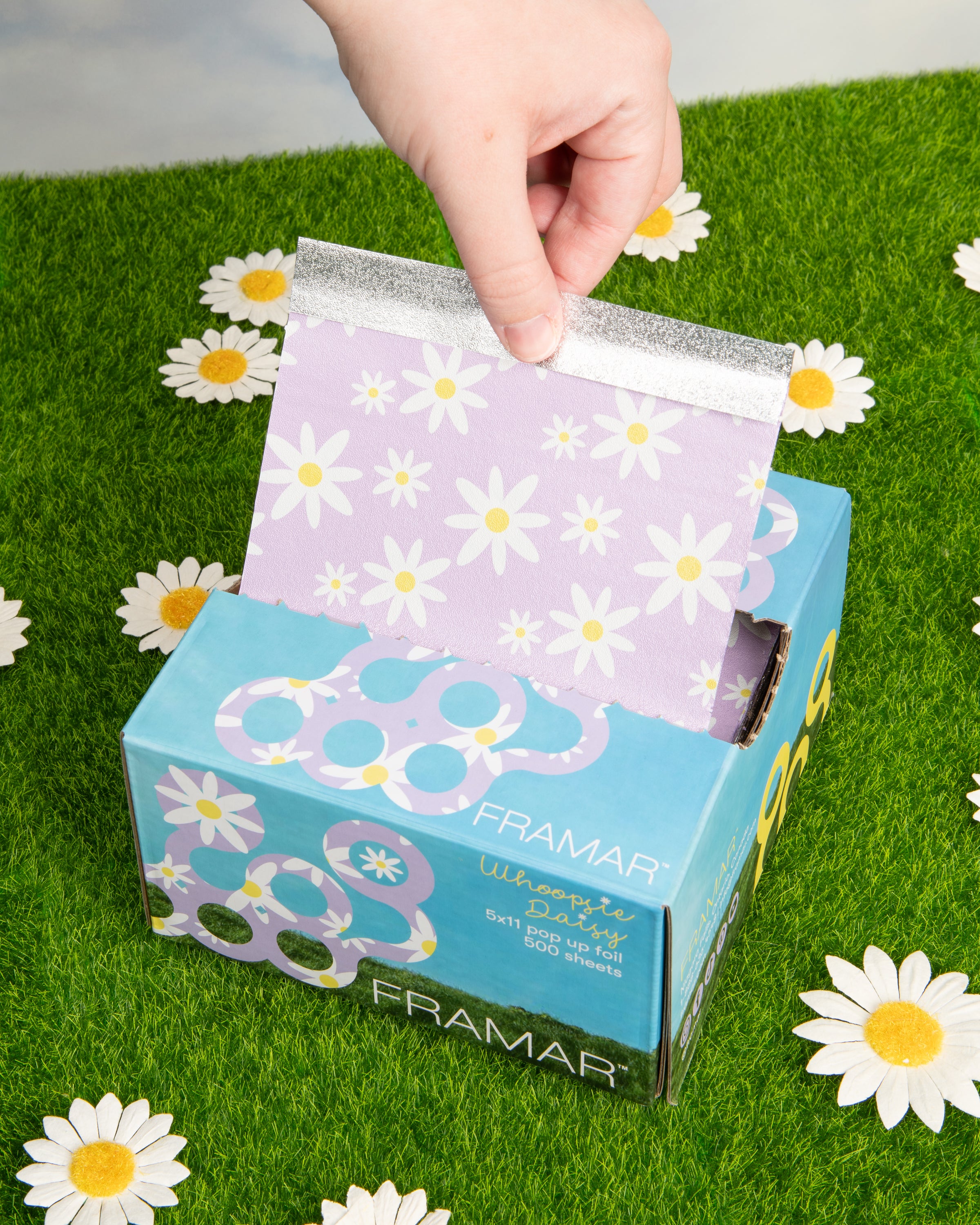 A hand pulling daisy-printed foil from a colorful Framar box on artificial grass with daisy decorations.