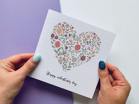 Hands holding a square card with flowers arranged in a heart shape on the front of it.