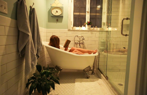 Woman sitting in a relaxing bath reading a book with candles on the window sill.