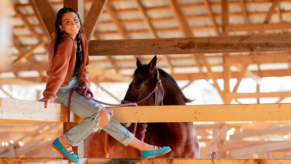 equestrian clothing women, young woman sitting on a fence holding a horse, while swinging her legs, wearing TuffRider GraphiX shoes.