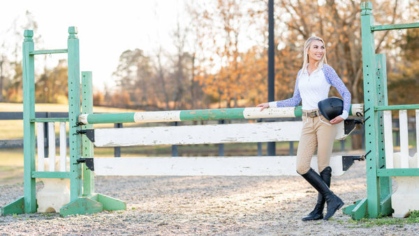 blonde woman wearing horse show accessories leans against a jump