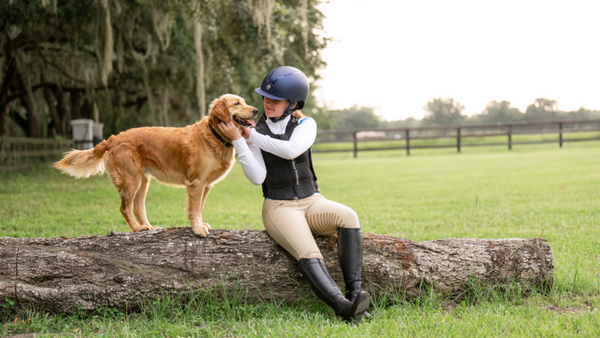 girl wearing horse riding safety gear sits on a log next to a golden retriever