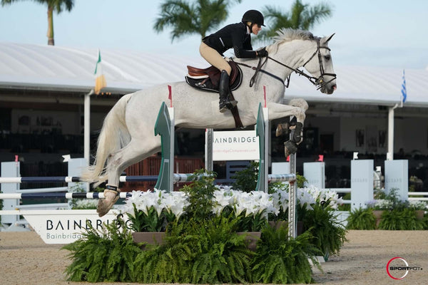 White horse and rider wearing a horse show outfit clear a large fence in the show jumping ring.