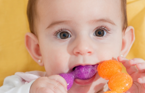 Bébé fille avec des boucles d'oreilles bébé et qui fait ses dents a un anneau de dentition en plastique orange et rose avec du gel réfrigérépère a mal aux dents et a la main de son père dans sa bouche. Son père a en main un anneau de dentition en bois comme ceux de Viwa