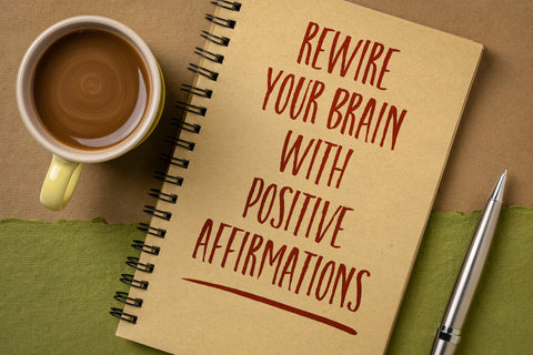 A table with a cup of coffee, a notebook and pen.  Notebook cover says rewire your brain with positive affirmations